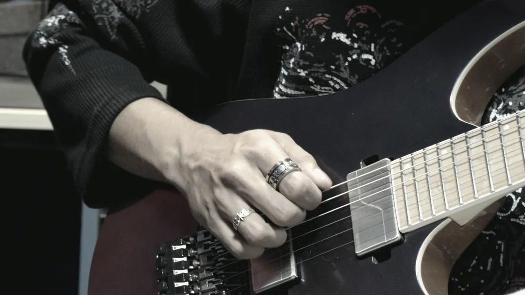 Close view of a player's hands over the pickups of an electric guitar, patterned sleeve resting against the body.