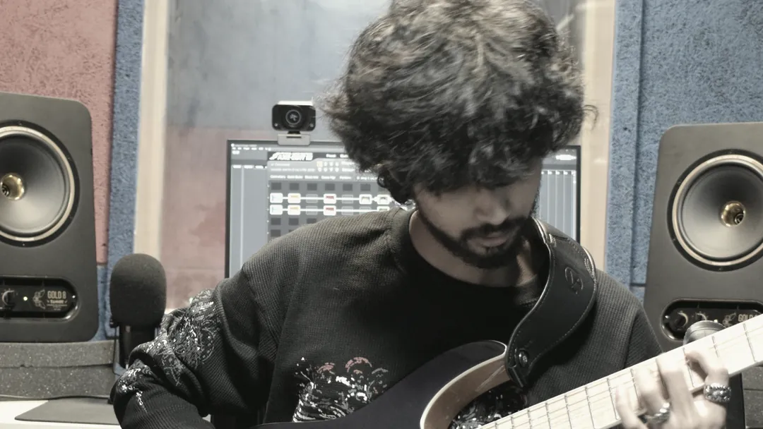 A guitarist seated at the desk working through a part, screen and monitors in front of them.